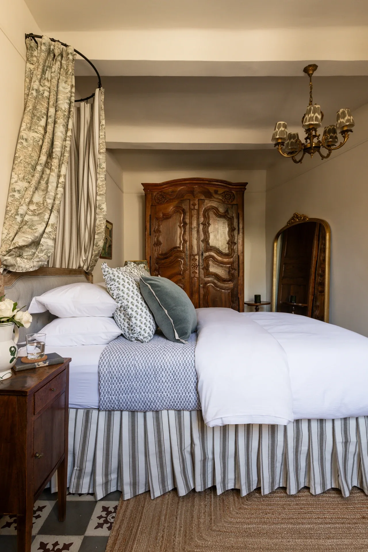 Chambre One at Maison de Frederic — a queen bed with its toile canopy drawn back on a curved rail, a carved walnut armoire and gilt mirror beyond, and a ticking-stripe bed skirt over patterned tiles